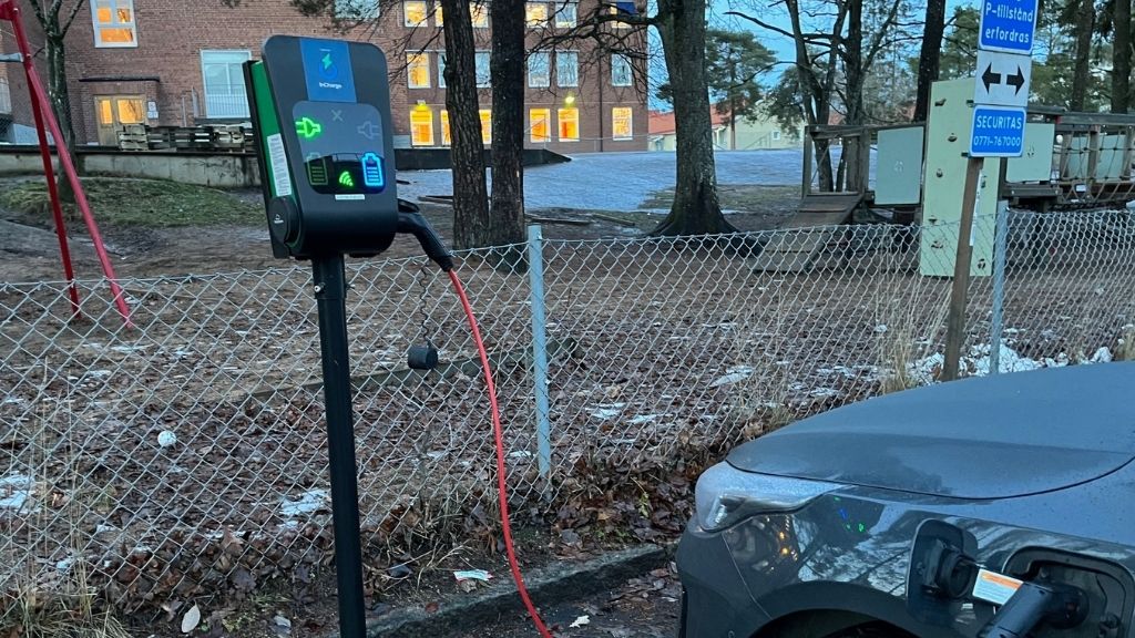 Ebil står parkerad och laddas av elstolpe som är framför ett staket. Till höger om bilen syns en parkeringsskylt.